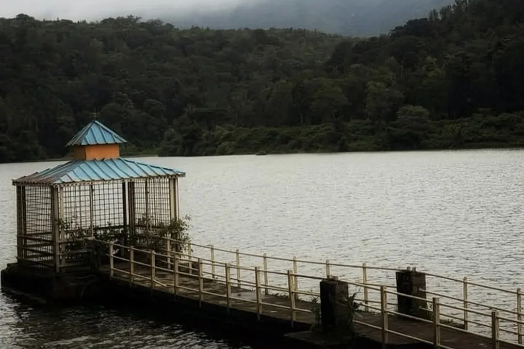 A railed walkway running out to a roofed intake pavilion standing in Hirekolale Lake, wooded hills and low cloud behind
